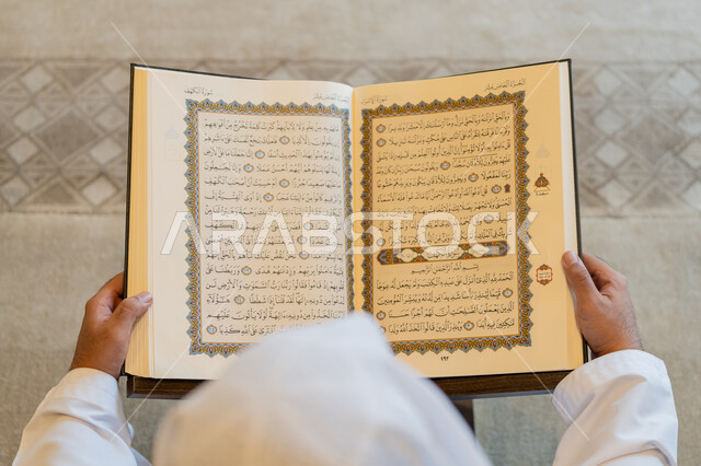العبادة والطاعة والتقرب الى الله,  تدبر الآيات القرآنية والتفكر بها, الالتزام بالورد اليومي,  صورة من الاعلى لرجل عربي خليجي إماراتي يرتدي الكندورة والغترة البيضاء يجلس في المسجد ويقرأ من المصحف الشريف، التمعن بسور القرآن الكريم، التعبد في بيوت الرحمن
