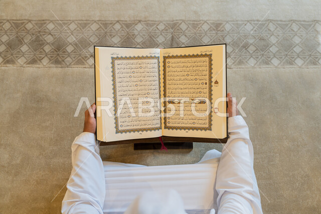 العبادة والطاعة والتقرب الى الله,  تدبر الآيات القرآنية والتفكر بها, الالتزام بالورد اليومي,  صورة من الاعلى لرجل عربي خليجي إماراتي يرتدي الكندورة والغترة البيضاء يجلس في المسجد ويقرأ من المصحف الشريف، التمعن بسور القرآن الكريم، التعبد في بيوت الرحمن