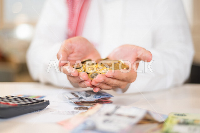 رجل عربي خليجي سعودي يحمل بيده كومة من النقود المعدنية، يعمل على ادارة الحسابات، النمو والأرباح المالية، إدخار وتوفير الاموال، عملات نقدية معدنية وورقية للريال السعودي، استخدام الالة الحاسبة لاجراء العمليات الحسابية