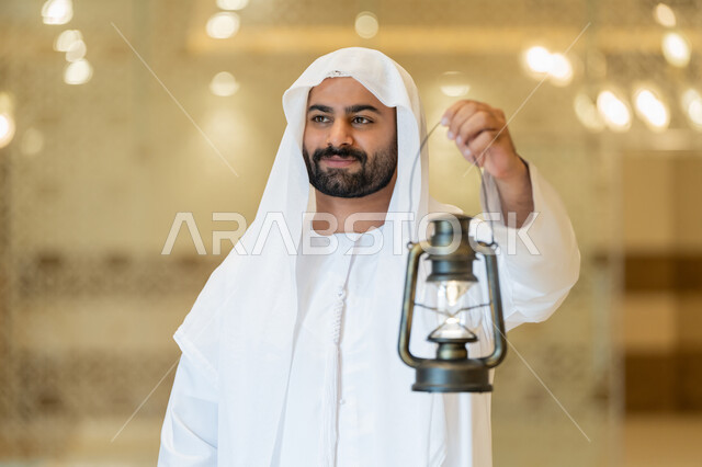 التصاميم والديكورات الداخلية للمساجد في الإمارات، مفهوم التقرب إلى الله، المناسبات الدينية والأعياد الإسلامية, الاستعداد لقدوم شهر رمضان المبارك, رجل عربي خليجي إماراتي يرتدي الكندورة والغترة البيضاء يقف في المسجد ويمسك بيده فانوس بإيماءات السعادة والسرور