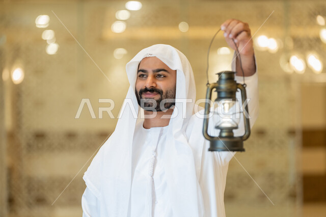العبادة والتقرب إلى الله، المناسبات الدينية والأعياد الإسلامية, الاستعداد لقدوم شهر رمضان المبارك, التصاميم والديكورات الداخلية للمساجد في الإمارات، رجل عربي خليجي إماراتي يرتدي الكندورة والغترة البيضاء يقف في المسجد ويمسك بيده فانوس بإيماءات السعادة والسرور