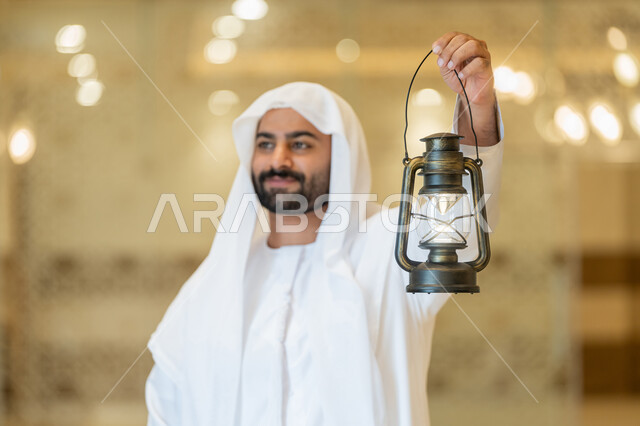 مفهوم التقرب إلى الله، المناسبات الدينية والأعياد الإسلامية, الاستعداد لقدوم شهر رمضان المبارك, التصاميم والديكورات الداخلية للمساجد في الإمارات، رجل عربي خليجي إماراتي يرتدي الكندورة والغترة البيضاء يقف في المسجد ويمسك بيده فانوس بإيماءات السعادة والسرور