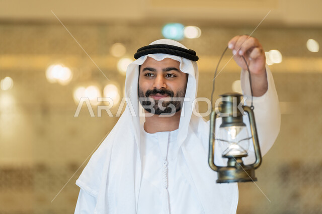 العبادة والتقرب إلى الله، المناسبات الدينية والأعياد الإسلامية, الاستعداد لقدوم شهر رمضان المبارك, التصاميم والديكورات الداخلية للمساجد في الإمارات، رجل عربي خليجي إماراتي يرتدي الكندورة والغترة البيضاء يقف في المسجد ويمسك بيده فانوس بإيماءات السعادة والسرور