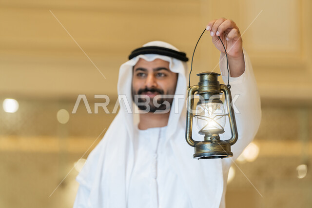 مفهوم التقرب إلى الله، المناسبات الدينية والأعياد الإسلامية, الاستعداد لقدوم شهر رمضان المبارك, التصاميم والديكورات الداخلية للمساجد في الإمارات، رجل عربي خليجي إماراتي يرتدي الكندورة والغترة البيضاء يقف في المسجد ويمسك بيده فانوس بإيماءات السعادة والسرور