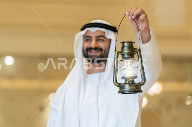 التصاميم والديكورات الداخلية للمساجد في الإمارات، مفهوم التقرب إلى الله، المناسبات الدينية والأعياد الإسلامية, الاستعداد لقدوم شهر رمضان المبارك, رجل عربي خليجي إماراتي يرتدي الكندورة والغترة البيضاء يقف في المسجد ويمسك بيده فانوس بإيماءات السعادة والسرور