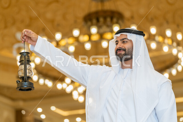 مفهوم التقرب إلى الله، المناسبات الدينية والأعياد الإسلامية, الاستعداد لقدوم شهر رمضان المبارك, التصاميم والديكورات الداخلية للمساجد في الإمارات، رجل عربي خليجي إماراتي يرتدي الكندورة والغترة البيضاء يقف في المسجد ويمسك بيده فانوس بإيماءات السعادة والسرور