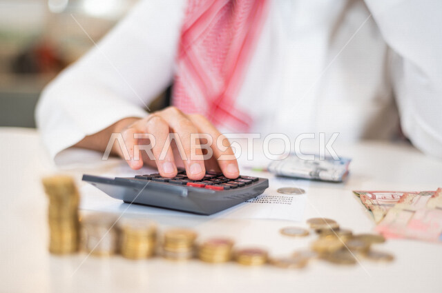رجل عربي خليجي سعودي يعمل على ادارة الحسابات، النمو والأرباح المالية، إدخار وتوفير الاموال، عملات نقدية معدنية وورقية للريال السعودي، استخدام الالة الحاسبة لاجراء العمليات الحسابية