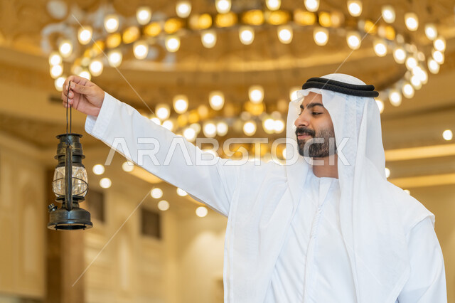 العبادة والتقرب إلى الله، المناسبات الدينية والأعياد الإسلامية, الاستعداد لقدوم شهر رمضان المبارك, التصاميم والديكورات الداخلية للمساجد في الإمارات، رجل عربي خليجي إماراتي يرتدي الكندورة والغترة البيضاء يقف في المسجد ويمسك بيده فانوس بإيماءات السعادة والسرور