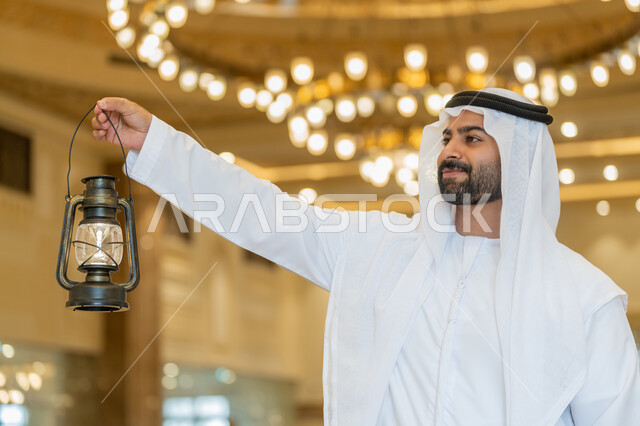 التصاميم والديكورات الداخلية للمساجد في الإمارات، الاحتفال بقدوم شهر رمضان المبارك, العبادة والتقرب إلى الله, أداء الفرائض الدينية, رجل عربي خليجي إماراتي يرتدي الكندورة والغترة البيضاء يقف في المسجد ويمسك بيده فانوس بإيماءات السعادة والسرور