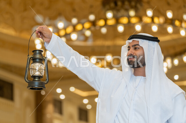 المناسبات الدينية والأعياد الإسلامية, الاستعداد لقدوم شهر رمضان المبارك, التصاميم والديكورات الداخلية للمساجد في الإمارات، العبادة والتقرب إلى الله، رجل عربي خليجي إماراتي يرتدي الكندورة والغترة البيضاء يقف في المسجد ويمسك بيده فانوس بإيماءات السعادة والسرور