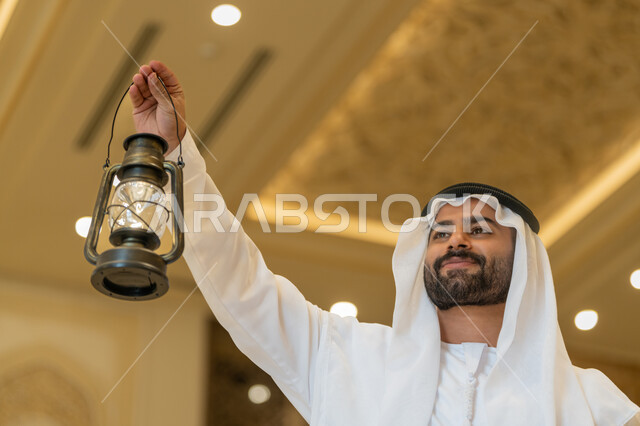 التصاميم والديكورات الداخلية للمساجد في الإمارات، الاحتفال بقدوم شهر رمضان المبارك, العبادة والتقرب إلى الله, أداء الفرائض الدينية, رجل عربي خليجي إماراتي يرتدي الكندورة والغترة البيضاء يقف في المسجد ويمسك بيده فانوس بإيماءات السعادة والسرور