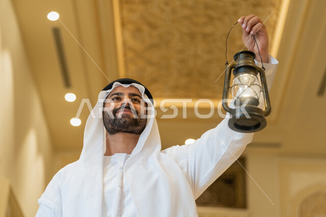 المناسبات الدينية والأعياد الإسلامية, الاستعداد لقدوم شهر رمضان المبارك, التصاميم والديكورات الداخلية للمساجد في الإمارات، العبادة والتقرب إلى الله، رجل عربي خليجي إماراتي يرتدي الكندورة والغترة البيضاء يقف في المسجد ويمسك بيده فانوس بإيماءات السعادة والسرور