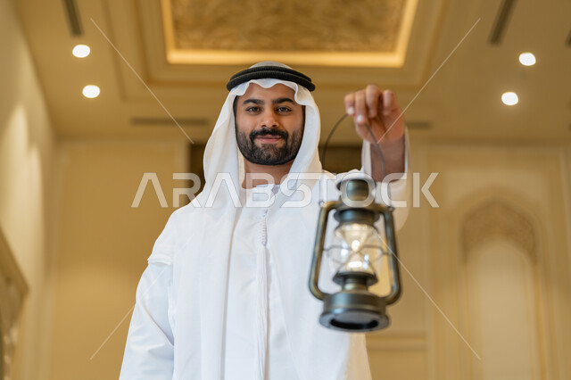 التصاميم والديكورات الداخلية للمساجد في الإمارات، الاحتفال بقدوم شهر رمضان المبارك, العبادة والتقرب إلى الله, أداء الفرائض الدينية, رجل عربي خليجي إماراتي يرتدي الكندورة والغترة البيضاء يقف في المسجد ويمسك بيده فانوس بإيماءات السعادة والسرور