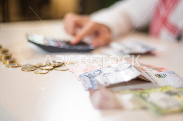 رجل عربي خليجي سعودي يعمل على ادارة الحسابات، النمو والأرباح المالية، إدخار وتوفير الاموال، عملات نقدية معدنية وورقية للريال السعودي، استخدام الالة الحاسبة لاجراء العمليات الحسابية