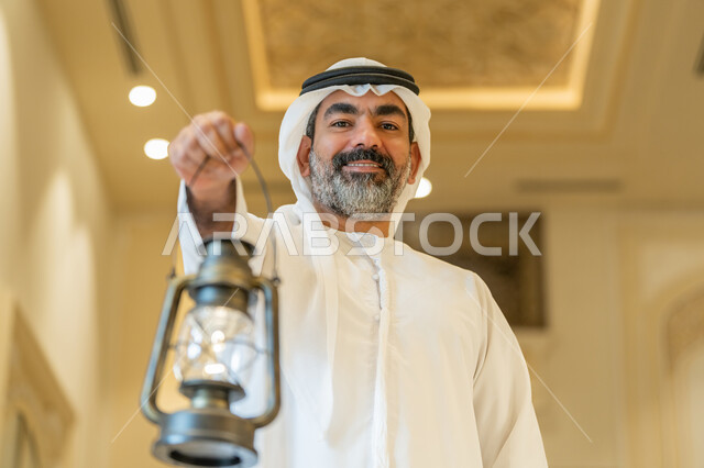 Celebrating the advent of the holy month of Ramadan, worship and getting closer to God, performing religious duties, interior designs and decorations of mosques in the Emirates, an Arab Gulf Emirati man wearing a kandora and a white ghutra standing in the mosque and holding a lantern in his hand with gestures of happiness and pleasure.