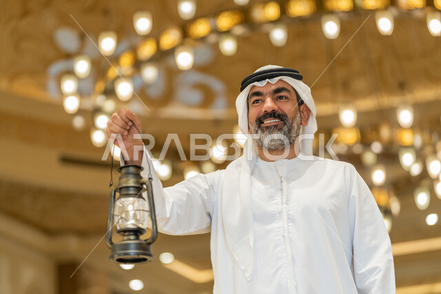 التصاميم والديكورات الداخلية للمساجد في الإمارات، الاحتفال بقدوم شهر رمضان المبارك, العبادة والتقرب إلى الله, أداء الفرائض الدينية,  رجل عربي خليجي إماراتي يرتدي الكندورة والغترة البيضاء يقف في المسجد ويمسك بيده فانوس بإيماءات السعادة والسرور