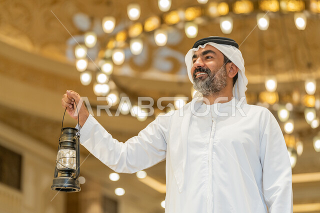 Celebrating the advent of the holy month of Ramadan, worship and getting closer to God, performing religious duties, interior designs and decorations of mosques in the Emirates, an Arab Gulf Emirati man wearing a kandora and a white ghutra standing in the mosque and holding a lantern in his hand with gestures of happiness and pleasure.