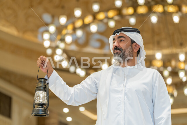 التصاميم والديكورات الداخلية للمساجد في الإمارات، الاحتفال بقدوم شهر رمضان المبارك, العبادة والتقرب إلى الله, أداء الفرائض الدينية,  رجل عربي خليجي إماراتي يرتدي الكندورة والغترة البيضاء يقف في المسجد ويمسك بيده فانوس بإيماءات السعادة والسرور