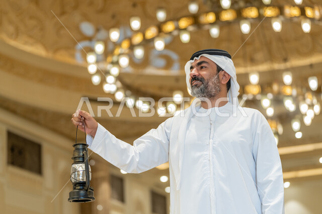 Worship and getting closer to God, interior designs and decorations of mosques in the Emirates, celebrating the advent of the holy month of Ramadan, performing religious duties, an Arab Gulf Emirati man wearing a kandora and a white ghutra standing in the mosque and holding a lantern in his hand with gestures of happiness and pleasure.