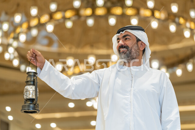 التصاميم والديكورات الداخلية للمساجد في الإمارات، الاحتفال بقدوم شهر رمضان المبارك, العبادة والتقرب إلى الله, أداء الفرائض الدينية,  رجل عربي خليجي إماراتي يرتدي الكندورة والغترة البيضاء يقف في المسجد ويمسك بيده فانوس بإيماءات السعادة والسرور