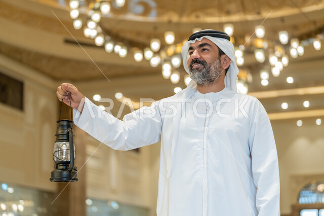 Celebrating the advent of the holy month of Ramadan, worship and getting closer to God, performing religious duties, interior designs and decorations of mosques in the Emirates, an Arab Gulf Emirati man wearing a kandora and a white ghutra standing in the mosque and holding a lantern in his hand with gestures of happiness and pleasure.