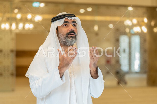 مفهوم الخشوع والتضرع, طلب الرحمة والمغفرة, العبادة والتقرب الى الله تعالى, رجل عربي خليجي اماراتي يرتدي الكندورة والغترة البيضاء يقف في إحدى المساجد ويرفع يديه للأعلى بتعابير الدعاء, أداء الفرائض الدينية الإسلامية، مفهوم أركان الإسلام