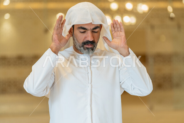 إقامة الصلاة على وقتها, العبادة والتقرب إلى الله، أداء الفرائض الدينية الإسلامية، الركن الثاني من أركان الإسلام، صورة مقربة لرجل عربي خليجي إماراتي يرتدي الكندورة والغترة البيضاء يقف باستقامة يرفع يديه لتكبيرة الإحرام, الشعور بالخشوع والطمأنينة