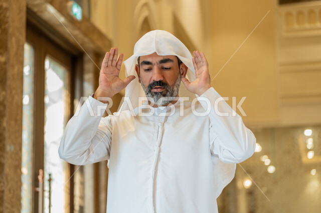 أداء الفرائض الدينية الإسلامية، إقامة الصلاة على وقتها, العبادة والتقرب إلى الله، الركن الثاني من أركان الإسلام، صورة مقربة لرجل عربي خليجي إماراتي يرتدي الكندورة والغترة البيضاء يقف باستقامة يرفع يديه لتكبيرة الإحرام, الشعور بالخشوع والطمأنينة