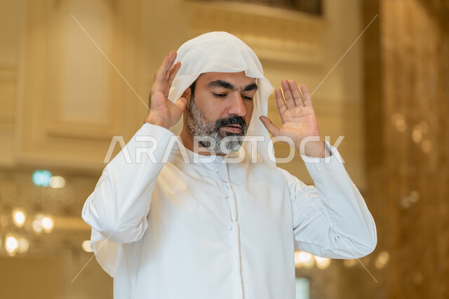 إقامة الصلاة على وقتها, العبادة والتقرب إلى الله، أداء الفرائض الدينية الإسلامية، الركن الثاني من أركان الإسلام، صورة مقربة لرجل عربي خليجي إماراتي يرتدي الكندورة والغترة البيضاء يقف باستقامة يرفع يديه لتكبيرة الإحرام, الشعور بالخشوع والطمأنينة