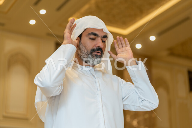أداء الفرائض الدينية الإسلامية، إقامة الصلاة على وقتها, العبادة والتقرب إلى الله، الركن الثاني من أركان الإسلام، صورة مقربة لرجل عربي خليجي إماراتي يرتدي الكندورة والغترة البيضاء يقف باستقامة يرفع يديه لتكبيرة الإحرام, الشعور بالخشوع والطمأنينة