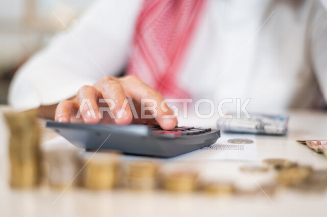 رجل عربي خليجي سعودي يعمل على ادارة الحسابات، النمو والأرباح المالية، إدخار وتوفير الاموال، عملات نقدية معدنية وورقية للريال السعودي، استخدام الالة الحاسبة لاجراء العمليات الحسابية