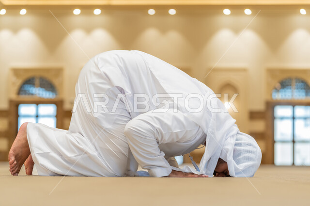 الركن الثاني من أركان الإسلام، الشعور بالخشوع والطمأنينة, إقامة الصلاة على وقتها, أداء الفرائض الدينية الإسلامية، العبادة والتقرب إلى الله، صورة من الجانب لرجل عربي خليجي إماراتي يرتدي الكندورة والغترة البيضاء يقوم بالسجود في احد المساجد