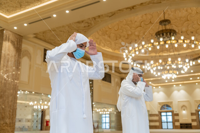 الحفاظ على التباعد، الالتزام باجراءات الوقاية، أداء فريضة الصلاه داخل المسجد، صلاة الجماعة لرجلان اماراتيان عربيان خليجيان يرتديان الكندورة والغترة البيضاء ويضعان الكمامة، العبادة و التقرب الى الله