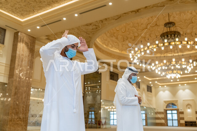 الحفاظ على التباعد، الالتزام باجراءات الوقاية، أداء فريضة الصلاه داخل المسجد، صلاة الجماعة لرجلان اماراتيان عربيان خليجيان يرتديان الكندورة والغترة البيضاء ويضعان الكمامة، العبادة و التقرب الى الله
