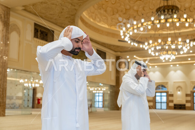 ايماءات التكبير لبدء الصلاة، أداء الفرائض بأوقاتها، رجلان عربيان خليجيان اماراتيان يرتديان الكندورة والغترة البيضاء يقفون على السجادة يقوم بالتكبير لأداء الصلاة، العبادة والتقرب إلى الله، الشعور بالخشوع والطمأنينة