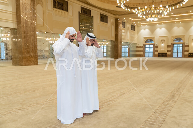 أداء الفرائض بأوقاتها، ايماءات التكبير لبدء الصلاة، رجلان عربيان خليجيان اماراتيان يرتديان الكندورة والغترة البيضاء يقفون على السجادة يقوم بالتكبير لأداء الصلاة، العبادة والتقرب إلى الله، الشعور بالخشوع والطمأنينة، الجسم بالطول الكامل