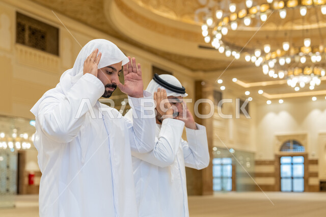 ايماءات التكبير لبدء الصلاة، أداء الفرائض بأوقاتها، رجلان عربيان خليجيان اماراتيان يرتديان الكندورة والغترة البيضاء يقفون على السجادة يقوم بالتكبير لأداء الصلاة، العبادة والتقرب إلى الله، الشعور بالخشوع والطمأنينة