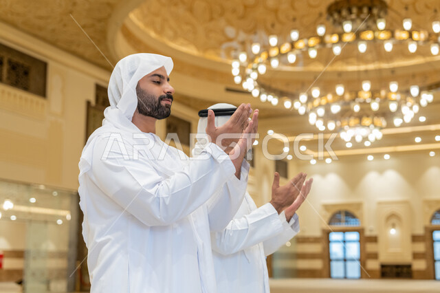مفهوم أركان الإسلام، أداء فرائض الصلاة بأوقاتها، الدعاء و التضرع لرب العباد، رجلان عربيان خليجيان اماراتيان يرتدون الكندورة والغترة البيضاء يقفون على السجادة بأيادي مفتوحة للأعلى، العبادة والتقرب إلى الله،