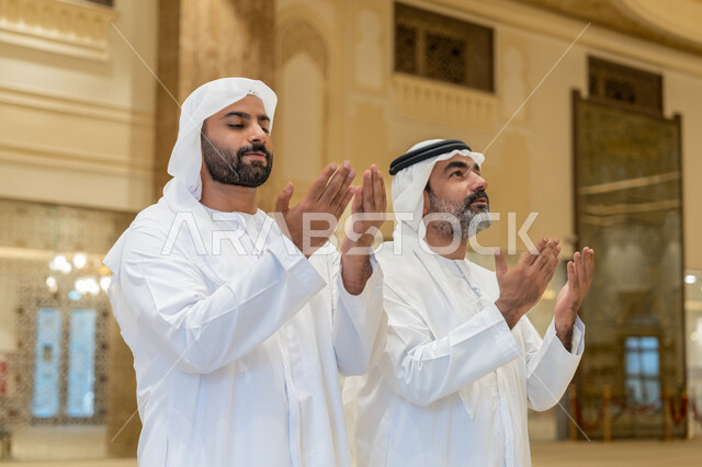 أداء فرائض الصلاة بأوقاتها، الدعاء و التضرع لرب العباد، مفهوم أركان الإسلام، رجلان عربيان خليجيان اماراتيان يرتدون الكندورة والغترة البيضاء يقفون على السجادة بأيادي مفتوحة للأعلى، العبادة والتقرب إلى الله،