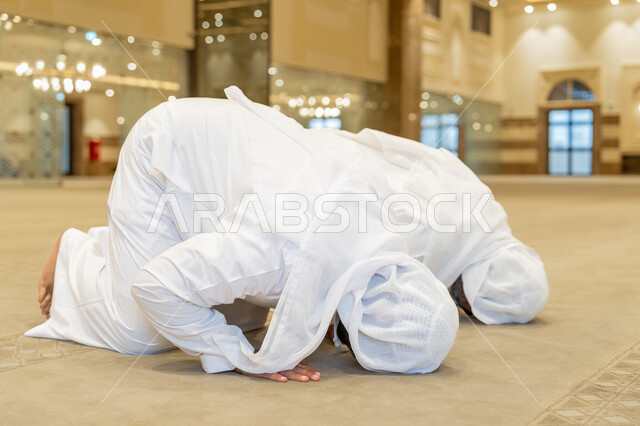 أداء الفرائض الدينية الإسلامية، الركن الثاني من أركان الإسلام، إقامة الصلاة على وقتها, العبادة والتقرب إلى الله، صورة مقربة لرجلين عربيين خليجيين إماراتيين يرتديين الكندورة والغترة البيضاء يقومان بالسجود, الشعور بالخشوع والطمأنينة