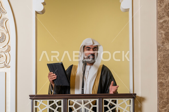 إلقاء خطاب و دروس توجيهية على المسلمين, توجيه الناس لطريق الحق والهداية, الحث على أداء العبادات والفرائض الدينية, إمام مسجد عربي خليجي إماراتي يرتدي البشت والكندورة والغترة البيضاء يقف على المنبر ويلقي خطبة يوم الجمعة من الجهاز اللوحي