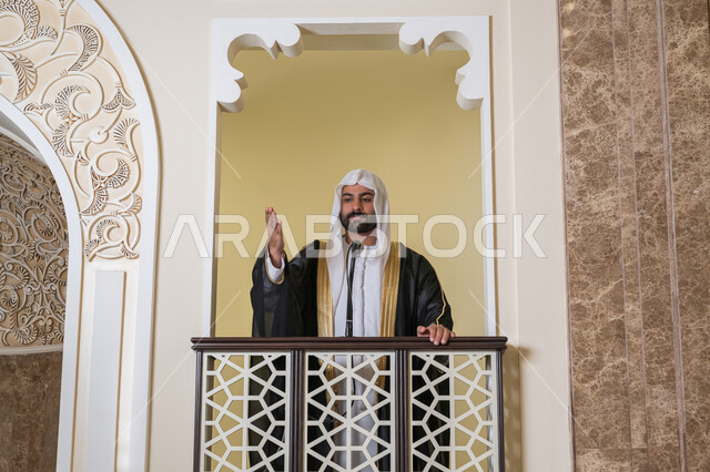 دور أئمة المساجد والخطباء في نشر الدين، مفهوم العبادة والتقرب إلى الله تعالى, توجيه الناس نحو الحق والهداية, إمام عربي خليجي إماراتي يرتدي البشت والكندورة والغترة البيضاء يقف أمام مكبر الصوت ويلقي الخطبة على المنبر, نداء المسلمين لأداء الفرائض