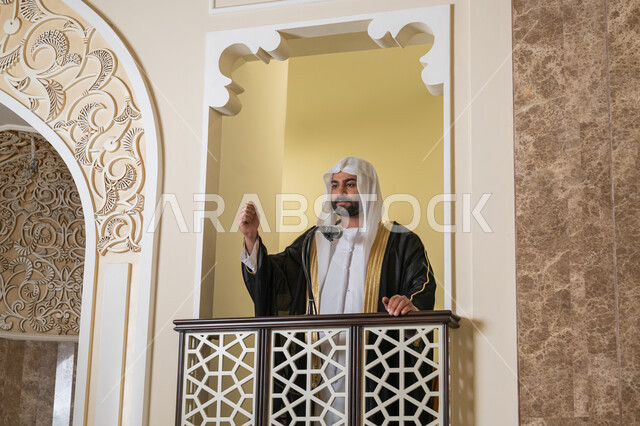 دور أئمة المساجد والخطباء في نشر الدين، مفهوم العبادة والتقرب إلى الله تعالى, توجيه الناس نحو الحق والهداية, إمام عربي خليجي إماراتي يرتدي البشت والكندورة والغترة البيضاء يقف أمام مكبر الصوت ويلقي الخطبة على المنبر, نداء المسلمين لأداء الفرائض