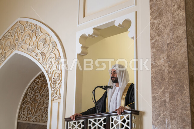 دور أئمة المساجد والخطباء في نشر الدين، مفهوم العبادة والتقرب إلى الله تعالى, توجيه الناس نحو الحق والهداية, إمام عربي خليجي إماراتي يرتدي البشت والكندورة والغترة البيضاء يقف أمام مكبر الصوت ويلقي الخطبة على المنبر, نداء المسلمين لأداء الفرائض