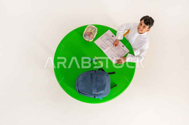 Developing artistic skills, enjoying leisure activities, practicing favorite hobbies during break time, looking at the camera with gestures of happiness and pleasure, vertical top portrait of a Saudi Arabian Gulf boy wearing traditional dress sitting at the table drawing and coloring, white background