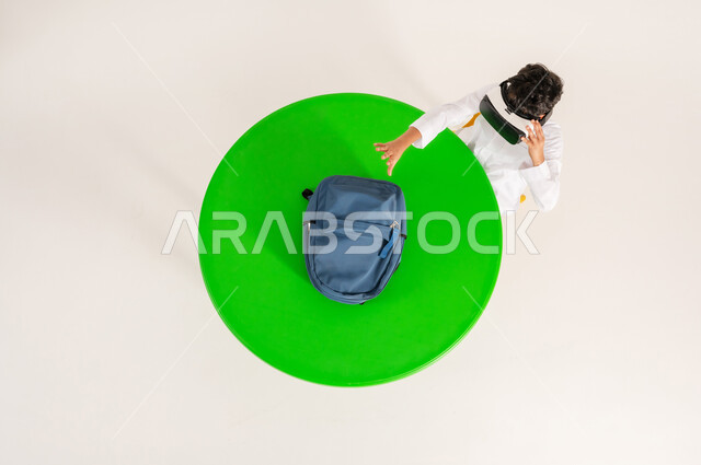 Using modern technologies in school education, interacting with the imaginary world, integrating virtual reality with learning and training, vertical top portrait of a Saudi Arabian Gulf boy wearing traditional dress, putting on 3D VR glasses and raising his hand to try to grab something, white background