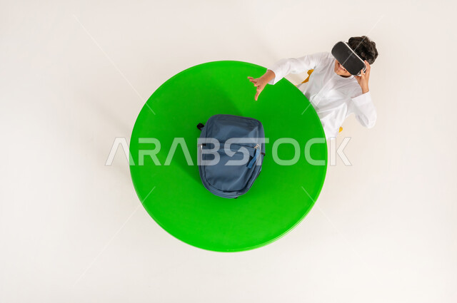 التفاعل مع العالم الخيالي, دمج الواقع الافتراضي بالتعلم والتدريب، استخدام التقنيات الحديثة في التعليم المدرسي, بورتريه علوي عامودي لولد عربي خليجي سعودي يرتدي الثوب التقليدي يضع نظارة الفي ار ثلاثية الأبعاد ويرفع يده لمحاولة الإمساك بشيء ما, خلفية بيضاء