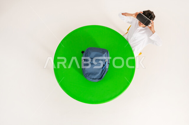 Integrating virtual reality with learning and training, the concept of simulating the imaginary world, using modern technologies in school education, vertical top portrait of a Saudi Arabian Gulf boy wearing traditional dress and putting on 3D VR glasses, white background