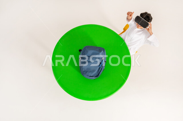 Concept of simulating the imaginary world, integrating virtual reality with learning and training, using modern technologies in school education, vertical top portrait of a Saudi Arabian Gulf boy wearing traditional dress, putting on 3D VR glasses and pointing his index finger up, white background