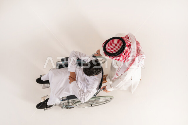 Family support and encouragement for people with disabilities, improving the lives of people with mobility disabilities, vertical top portrait of a Saudi Arabian Gulf man wearing traditional thobe helping his son with special needs, white background